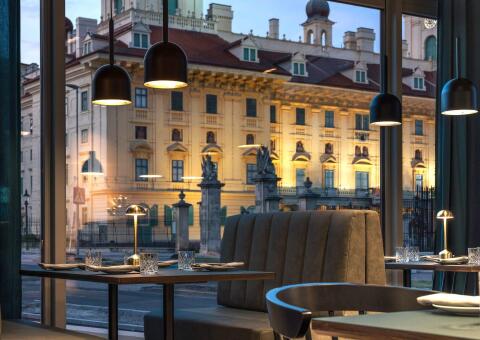 Blick aus einem modern eingerichteten Restaurant durch eine große Fensterfront auf ein prachtvolles, beleuchtetes historisches Gebäude in der Abenddämmerung. Im Vordergrund sind dunkel eingedeckte Tische mit Gläsern und kleinen goldenen Tischlampen zu sehen. Von der Decke hängen schlichte, schwarze Halbkugel-Leuchten, die ein warmes Licht abgeben. Eine gepolsterte Sitzbank und moderne Stühle laden zum Verweilen ein. Die Szenerie verbindet zeitgenössisches Design mit klassischer Architektur.