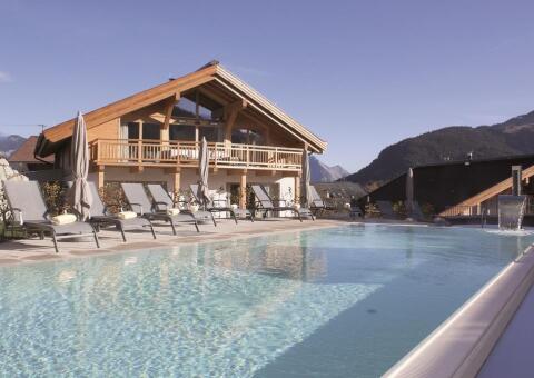 Blick auf einen eleganten Außenpool mit klarem, hellem Wasser und Schwalbenschwanz-Pooldusche. Am Beckenrand stehen zahlreiche graue Sonnenliegen mit Sonnenschirmen vor einem modernen, alpinen Holzchalet mit großem Balkon. Im Hintergrund erstreckt sich eine Berglandschaft unter klarem Himmel.