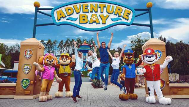 Eine vierköpfige Familie posiert jubelnd vor dem Eingang der „Adventure Bay“ im Movie Park Germany. Sie stehen unter einem großen blauen Torbogen mit der Aufschrift „Adventure Bay“ und springen mit erhobenen Armen in die Luft. Flankiert werden sie von vier lebensgroßen Maskottchen der PAW Patrol: Skye, Rubble, Chase und Marshall. Im Hintergrund ist der zentrale Aussichtsturm der PAW Patrol und ein bewaldeter Bereich unter blauem Himmel zu sehen.