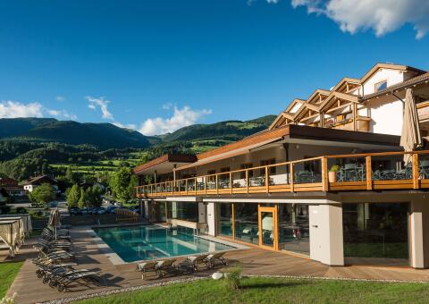 Ein modernes Wellnesshotel in den Alpen bei sonnigem Wetter. Im Vordergrund befindet sich ein rechteckiger Außenpool, umgeben von einer Holzterrasse mit zahlreichen Sonnenliegen. Das Hotelgebäude verfügt über eine großzügige Terrasse im ersten Stock mit Holzgeländer und bodentiefen Glasfronten im Erdgeschoss. Im Hintergrund erstrecken sich saftig grüne Berghänge unter einem klaren blauen Himmel mit vereinzelten weißen Wolken.