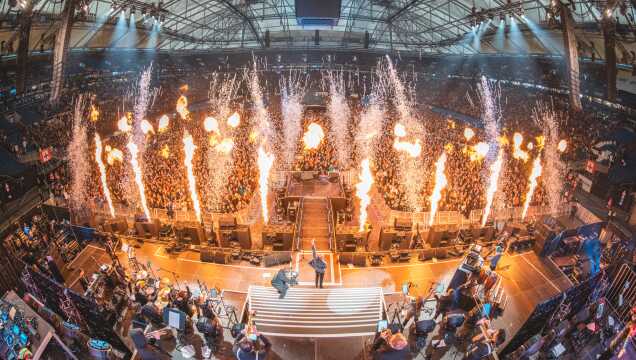 Spektakuläre Weitwinkelaufnahme eines Live-Konzerts in einem vollbesetzten Stadion. Auf der Bühne und inmitten der Zuschauermenge schießen hohe Flammenwerfer und Funkenfontänen empor. Die Arena ist hell erleuchtet, und im Vordergrund ist die technische Bühnenkonstruktion sowie ein Teil des Orchesters oder der Band zu sehen.