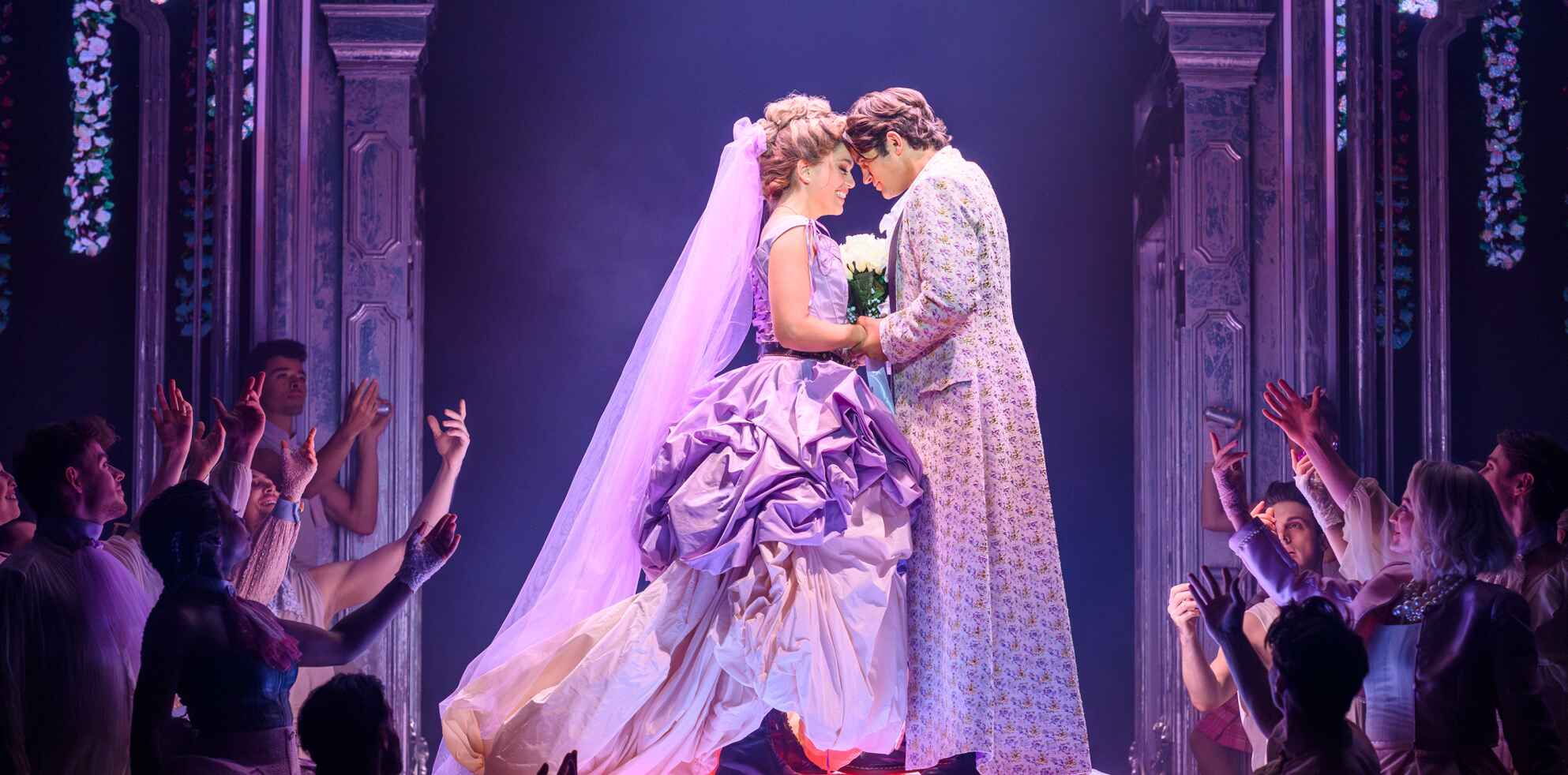 Braut und Bräutigam in eleganter Hochzeitskleidung stehen sich auf einer Theaterbühne innig gegenüber und berühren sich an der Stirn, umgeben von applaudierenden Darstellern unter violettem Bühnenlicht.