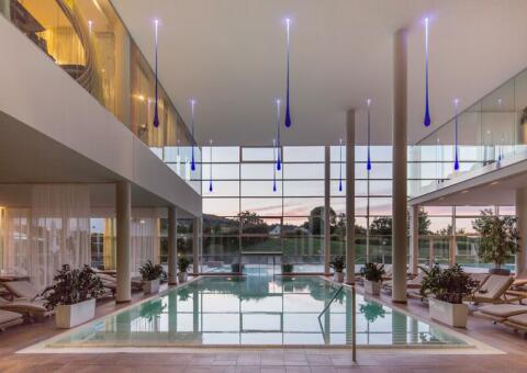 Ein moderner Hotel-Innenpool mit großen Glasfronten, die den Blick auf eine Hügellandschaft in der Abenddämmerung freigeben. Von der hohen Decke hängen kunstvolle, blau leuchtende Lichtelemente in Tropfenform herab. Links und rechts vom Becken stehen beigefarbene Liegen und weiße Pflanzkübel auf einem hellen Fliesenboden. Das Gebäude ist offen und großzügig über zwei Etagen gestaltet.