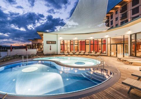 Ein beleuchteter Außenpoolbereich in der Abenddämmerung. Das geschwungene Becken ist in hell- und dunkelblaue Zonen unterteilt und wird von innen heraus angestrahlt. Dahinter befindet sich ein modernes, einstöckiges Hotelgebäude mit bodentiefen Glasfronten, durch die rote Vorhänge und eine warme Innenbeleuchtung schimmern. Ein großes, weißes Sonnensegel überspannt Teile der Terrasse, auf der zahlreiche beigefarbene Sonnenliegen bereitstehen. Im Hintergrund ist ein bewölkter Abendhimmel über einer dunklen Hügellandschaft zu sehen.