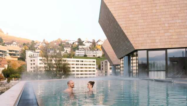 Blick von einer Terrasse mit lila Blumen im Vordergrund auf ein Thermal-Außenbecken. Die Anlage liegt eingebettet in ein grünes Tal mit bewaldeten Hängen und einer steilen Felswand im Hintergrund. Ein Wasserfall stürzt an der gegenüberliegenden Bergseite in die Tiefe.