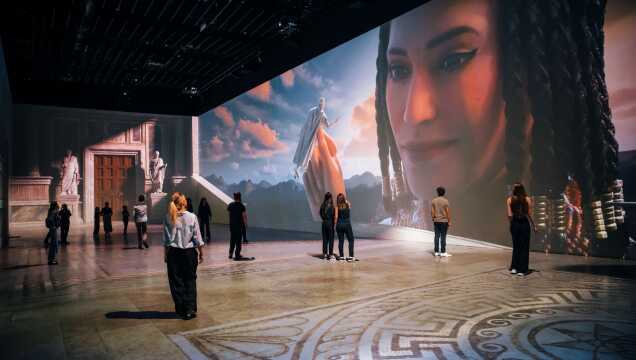 Eine immersive Multimedia-Ausstellung in einem großen Saal. Auf die Wände werden riesige Projektionen geworfen, die eine antike Architektur mit Statuen sowie ein monumentales Porträt einer Frau vor einer Berglandschaft bei Sonnenuntergang zeigen. Mehrere Besucher stehen im Raum und betrachten die Projektionen; der Boden ist mit einem klassischen Mosaikmuster verziert.