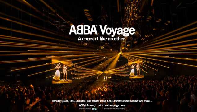 Blick in die vollbesetzte ABBA Arena in London während der virtuellen „ABBA Voyage“ Show. Unzählige goldene Laserstrahlen durchschneiden den abgedunkelten Raum über den Köpfen des Publikums. Auf großen Bildschirmen im Hintergrund sind die digitalen Avatare der Bandmitglieder in glitzernden Kostümen zu sehen. Der Schriftzug „ABBA Voyage – A concert like no other“ sowie Songtitel sind als Text eingeblendet.