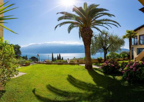 Blick von einem gepflegten Garten mit Rasenfläche und einer großen Palme auf einen tiefblauen See. Die Sonne scheint hell am klaren Himmel und wirft Schatten auf das Gras. Im Hintergrund erheben sich sanfte, wolkenverhangene Berge. Rechts sind ein mediterranes Gebäude und blühende Hortensienbüsche zu sehen.