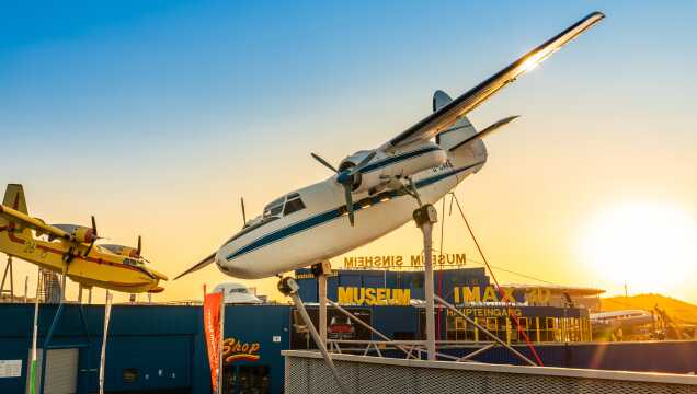 Blick auf das Außengelände des Museums bei Sonnenuntergang. Ein weiß-blaues Propellerflugzeug und ein gelbes Löschflugzeug sind auf hohen Stützen in Startposition aufgebockt. Im Hintergrund ist das Museumsgebäude mit der Aufschrift „MUSEUM SINSHEIM“ und dem „IMAX 3D“-Kino unter einem warmen Abendhimmel zu sehen.