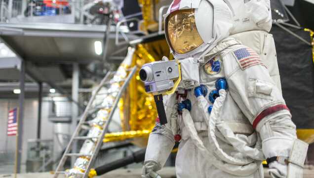 Ein Astronaut in einem weißen Raumanzug mit US-Flagge auf dem Oberarm und einer Kamera vor der Brust steht im Technikmuseum Speyer. Im Hintergrund sind Teile einer goldenen Mondlandefähre und eine Leiter zu sehen.